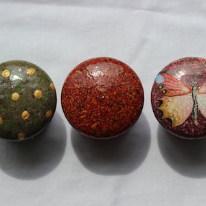 Könnte beinhalten: Drei dekorative Schrankknöpfe. Der erste ist olivgrün mit goldenen Punkten. Der zweite ist gesprenkelt rot. Der dritte ist bordeauxfarben mit einem Schmetterlingsdesign in Rot, Gelb und Blau.