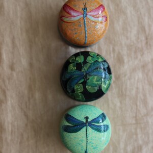 Może przedstawiać: Trzy okrągłe, malowane, ozdobne ozdoby z motywami ważek. G&oacute;rna ozdoba jest pomarańczowa z ważką w kolorze czerwonym i białym. Środkowa ozdoba jest czarna z ważką w kolorze zielonym i niebieskim. Dolna ozdoba jest jasnozielona z ważką w kolorze niebieskim.