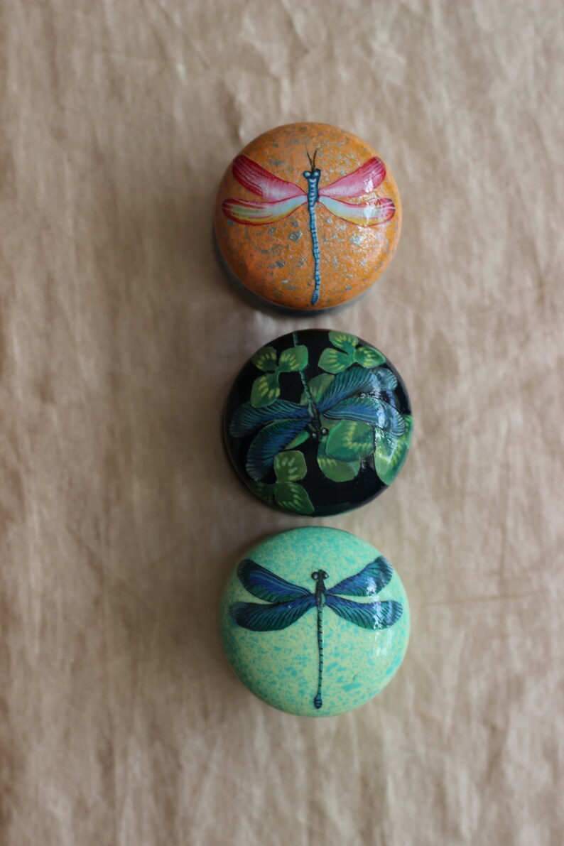 Może przedstawiać: Trzy okrągłe, malowane, ozdobne ozdoby z motywami ważek. G&oacute;rna ozdoba jest pomarańczowa z ważką w kolorze czerwonym i białym. Środkowa ozdoba jest czarna z ważką w kolorze zielonym i niebieskim. Dolna ozdoba jest jasnozielona z ważką w kolorze niebieskim.