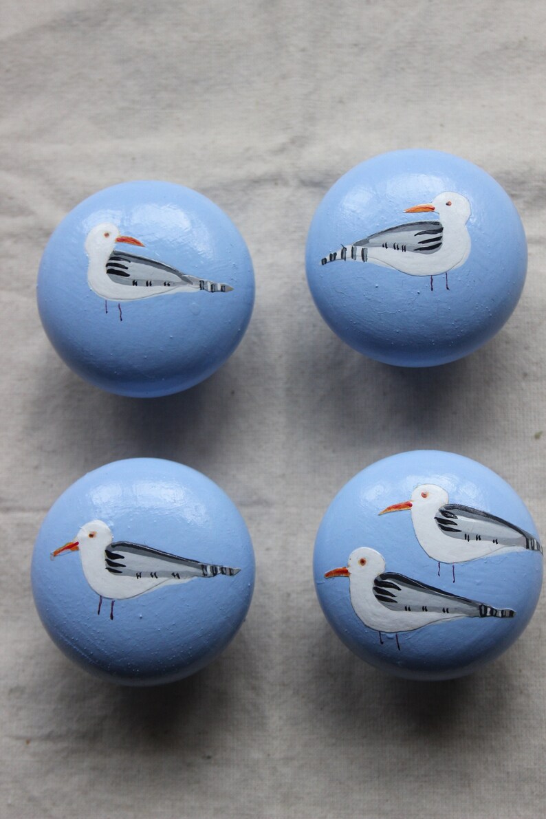 May include: Four light blue round wooden knobs with painted seagull designs. Each knob features a white seagull with black markings and an orange beak.