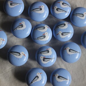 May include: A set of 16 blue painted wooden knobs with white seagull designs. Each knob features a pair of seagulls with grey wings and orange beaks.