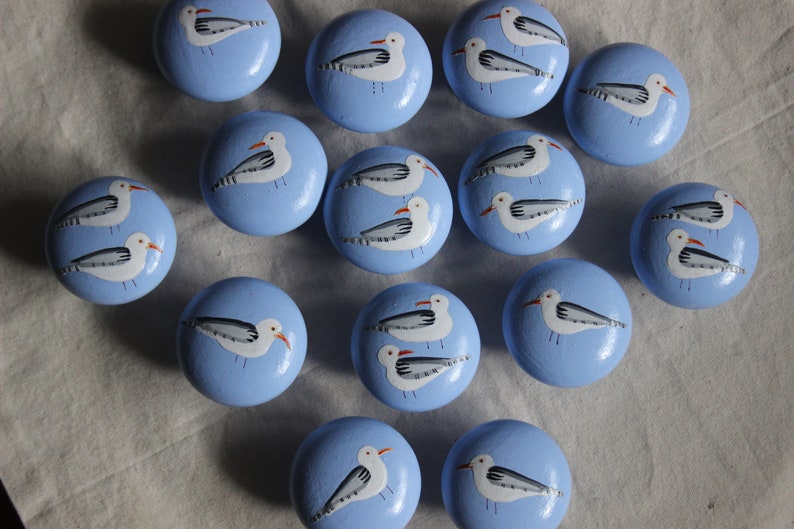 May include: A set of 16 blue painted wooden knobs with white seagull designs. Each knob features a pair of seagulls with grey wings and orange beaks.