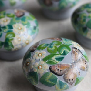 K&ouml;nnte beinhalten: F&uuml;nf runde, graue Keramik-Schmuckk&auml;stchen mit bemalten Blumen- und Schmetterlingsmotiven. Die K&auml;stchen sind mit gr&uuml;nen Bl&auml;ttern, wei&szlig;en Bl&uuml;ten und braunen Schmetterlingen verziert.