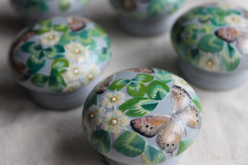 K&ouml;nnte beinhalten: F&uuml;nf runde, graue Keramik-Schmuckk&auml;stchen mit bemalten Blumen- und Schmetterlingsmotiven. Die K&auml;stchen sind mit gr&uuml;nen Bl&auml;ttern, wei&szlig;en Bl&uuml;ten und braunen Schmetterlingen verziert.