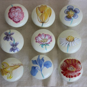 Może przedstawiać: Dziewięć okrągłych, kremowych gałek z malowanymi wzorami kwiatowymi. Wzory obejmują różową różę, żółty tulipan, fioletową bratkę, fioletową fiołkę, różowo-biały mak, białą stokrotkę, niebieski irys, czerwony mak i żółty narcyz.