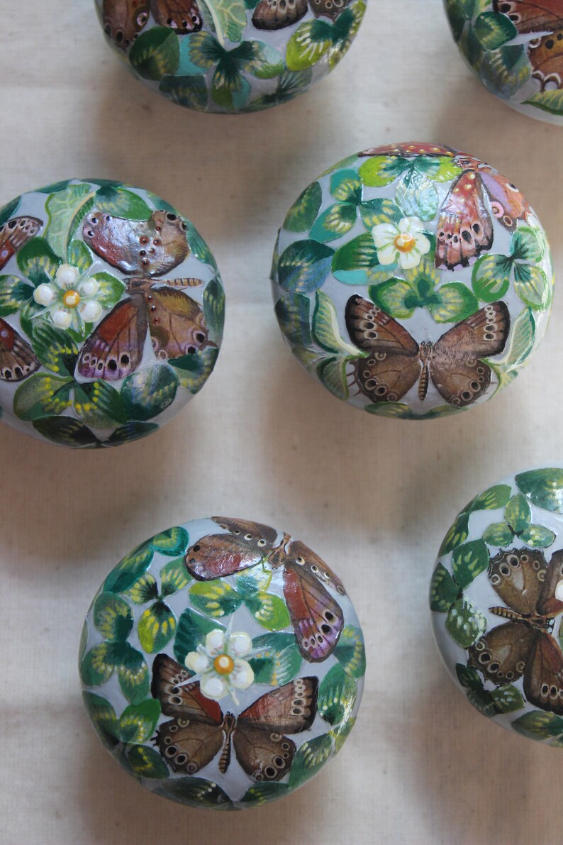 Puede incluir: Seis adornos redondos pintados con fondo azul claro. Cada adorno presenta un dise&ntilde;o de mariposa diferente con alas marrones, naranjas y blancas. Las mariposas est&aacute;n rodeadas de hojas verdes de tr&eacute;bol.