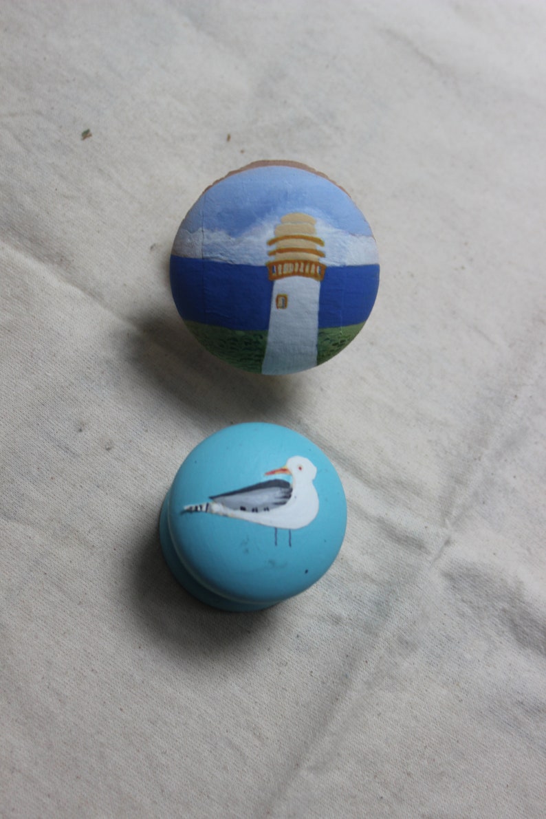 May include: Two hand-painted wooden knobs. One features a lighthouse scene with blue sky and sea. The other is light blue with a white seagull design. These knobs are perfect for adding a coastal touch to furniture.