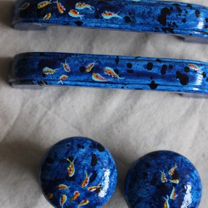 May include: Three blue painted drawer pulls with a design of small yellow and orange fish. The pulls are in the shape of a rectangle with rounded edges. Two of the pulls are long and one is round.