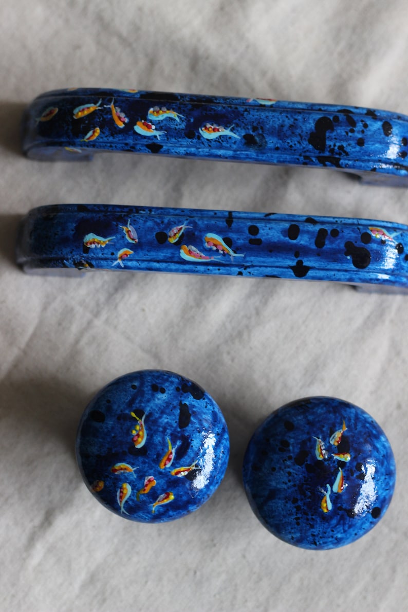 May include: Three blue painted drawer pulls with a design of small yellow and orange fish. The pulls are in the shape of a rectangle with rounded edges. Two of the pulls are long and one is round.