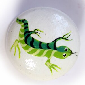 May include: A white, rounded stone features a hand-painted lizard design. The lizard is depicted in shades of green and black, with a striped body and detailed legs. The artwork is a whimsical, decorative item.
