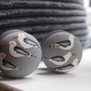 Pode incluir: Dois objetos decorativos redondos cinzentos com gaivotas pintadas à mão. Cada gaivota tem penas brancas, asas cinzentas e um bico laranja. Os objetos estão sobre uma superfície branca, com uma almofada cinzenta texturizada no fundo.