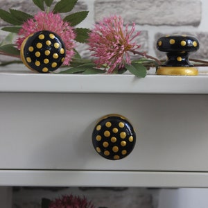 May include: Black and gold polka dot drawer knobs and a matching cabinet knob. The knobs are on a white drawer and cabinet. Pink flowers and green leaves are in the background.