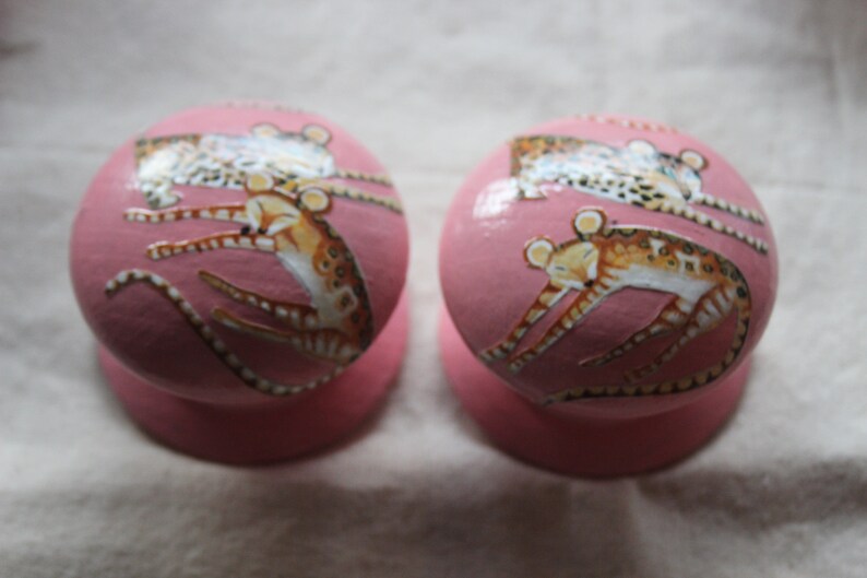Peut inclure: Deux boutons ronds roses avec une base blanche. Chaque bouton pr&eacute;sente un motif peint &agrave; la main d'un animal brun et blanc avec une queue enroul&eacute;e.