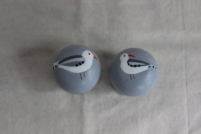 K&ouml;nnte beinhalten: Zwei runde, graue Keramikornamente mit bemalten M&ouml;wenmotiven. Die M&ouml;wen sind wei&szlig; mit schwarzen Markierungen und orangefarbenen Schn&auml;beln.