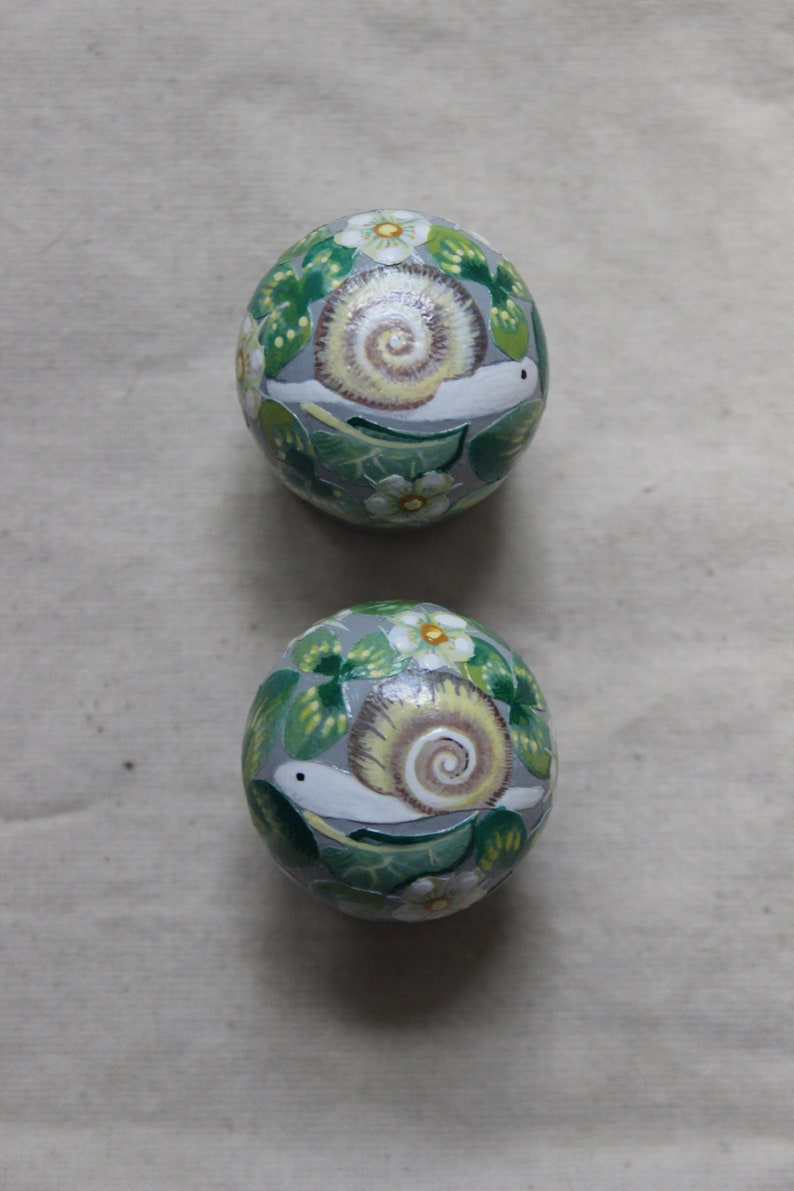 May include: Two decorative, hand-painted eggs. Each egg features a snail with a cream-colored shell and a white body, surrounded by green leaves and white flowers. The background of the eggs is a light gray color.