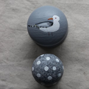 May include: Two grey painted wooden ornaments. One ornament features a white seagull with black stripes and a red beak. The other ornament is covered in white polka dots.