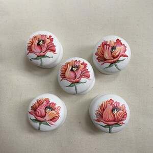 Puede incluir: Cinco pomos de armario blancos con un dise&ntilde;o floral pintado a mano. Cada pomo presenta una vibrante flor roja y naranja con hojas verdes. Los pomos son redondos y tienen una peque&ntilde;a base curva.