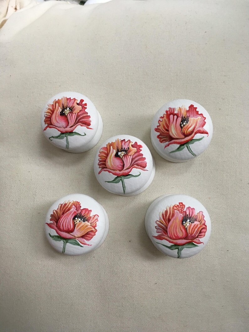 Puede incluir: Cinco pomos de armario blancos con un dise&ntilde;o floral pintado a mano. Cada pomo presenta una vibrante flor roja y naranja con hojas verdes. Los pomos son redondos y tienen una peque&ntilde;a base curva.