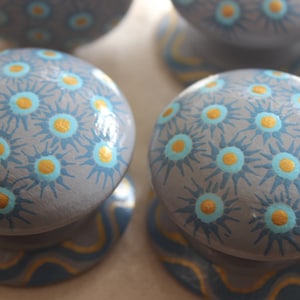 Puede incluir: Cuatro pomos de madera pintados de gris con un patrón de estallido de estrellas azul y dorado. Los pomos tienen una base ondulada azul y dorada.