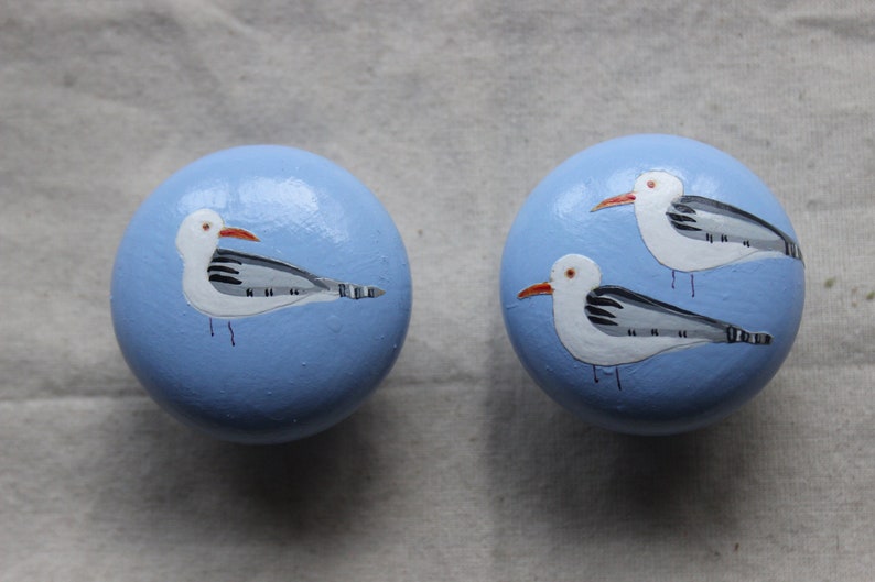 May include: Two light blue round wooden knobs with painted seagulls. Each knob features a white seagull with black and grey markings and a red beak.