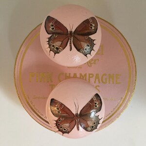 Peut inclure: Deux boutons d&eacute;coratifs ronds roses avec un motif de papillon. Les papillons ont des ailes marron avec des motifs d&eacute;taill&eacute;s. Les boutons sont sur une base rose circulaire avec le texte dor&eacute; "PINK CHAMPAGNE".