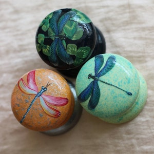 Puede incluir: Tres pomos redondos de madera con diseños de libélulas pintadas. Un pomo es negro con una libélula verde y azul, otro es verde claro con una libélula azul y el último es naranja con una libélula rosa y amarilla.
