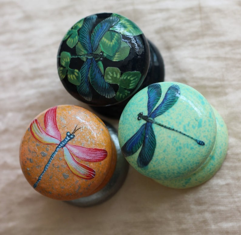 Może przedstawiać: Trzy okrągłe drewniane gałki z malowanymi wzorami ważek. Jedna gałka jest czarna z zielono-niebieską ważką, druga jest jasnozielona z niebieską ważką, a trzecia jest pomarańczowa z r&oacute;żowo-ż&oacute;łtą ważką.