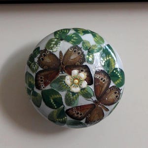 Könnte beinhalten: Ein rundes, dekoratives Objekt mit einem bemalten Schmetterlings- und Blumendesign. Die Schmetterlinge sind braun mit komplizierten Flügelmustern, vor einem weißen Hintergrund mit grünen Blättern und einer kleinen weißen Blume.