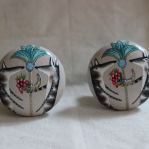 May include: Two round, hand-painted decorative objects. Each features a central design of a stylized blue and gold plant, flanked by two wolf-like figures. A horn of plenty and red berries complete the design.