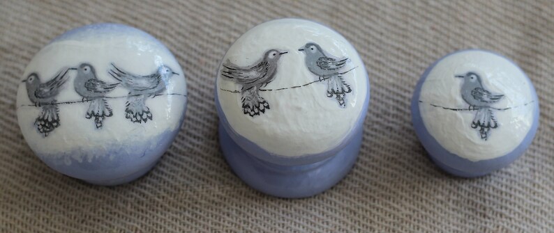 May include: A set of three ceramic knobs with a bird design. The knobs are white with a pale blue base and feature illustrations of birds perched on a wire. The knobs vary in size, suitable for drawers or cabinets.