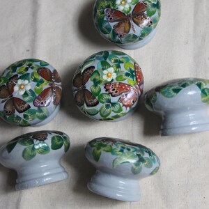 Puede incluir: Seis pomos de caj&oacute;n de cer&aacute;mica gris con un dise&ntilde;o pintado de hojas verdes, flores blancas y mariposas marrones.