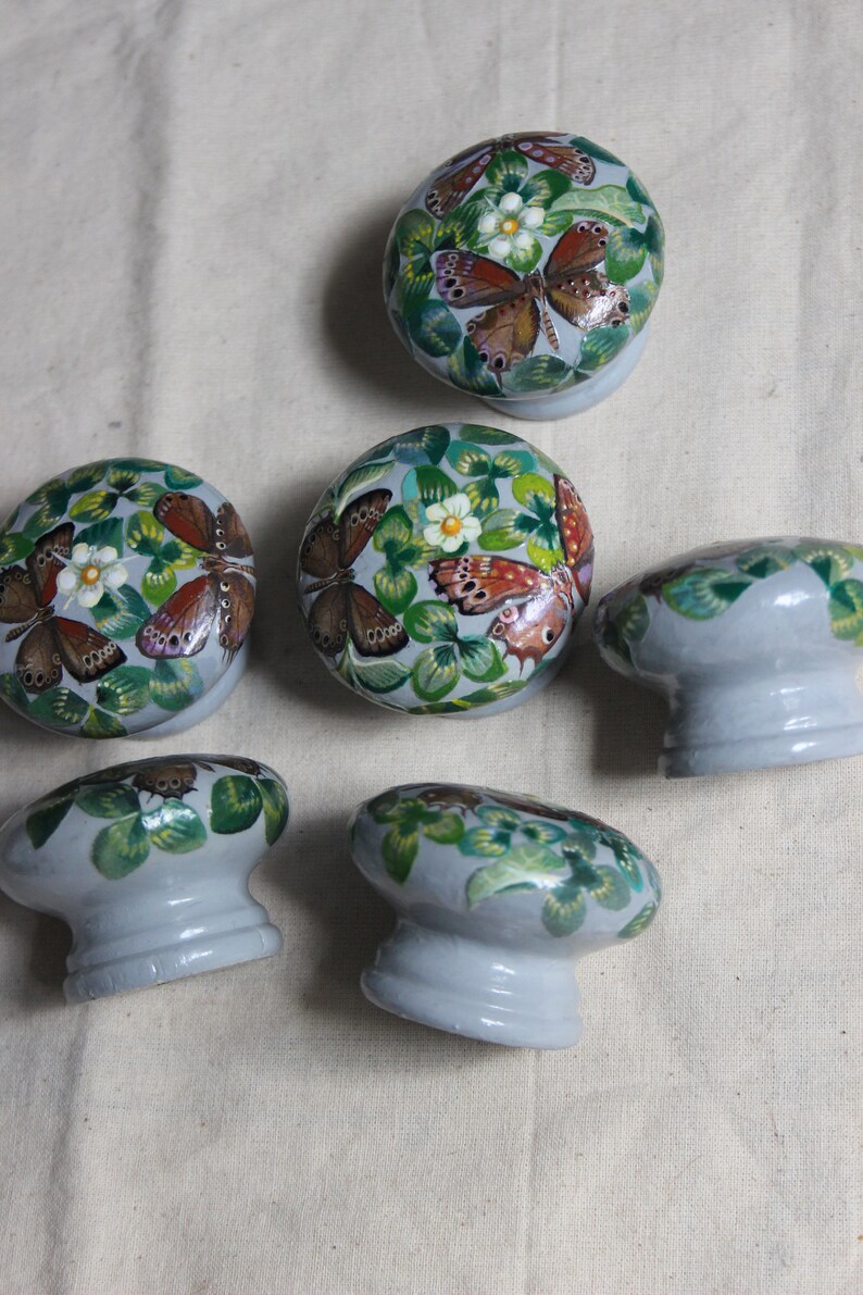 Puede incluir: Seis pomos de caj&oacute;n de cer&aacute;mica gris con un dise&ntilde;o pintado de hojas verdes, flores blancas y mariposas marrones.