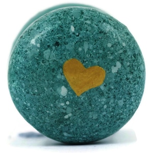 Op de afbeelding: Een turquoise gekleurde, gespikkelde steen met een gouden hart op het oppervlak. De steen heeft een afgeronde vorm en een textuur. Het gouden hart is gecentreerd op de steen.