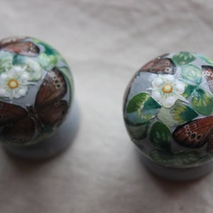 K&ouml;nnte beinhalten: Zwei runde Ornamente mit wei&szlig;em Hintergrund und gemalten Schmetterlingen und Blumen. Die Schmetterlinge sind braun mit schwarzen Markierungen und die Blumen sind wei&szlig; mit gelben Zentren.