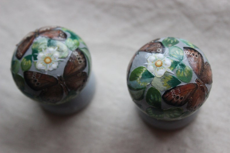 K&ouml;nnte beinhalten: Zwei runde Ornamente mit wei&szlig;em Hintergrund und gemalten Schmetterlingen und Blumen. Die Schmetterlinge sind braun mit schwarzen Markierungen und die Blumen sind wei&szlig; mit gelben Zentren.