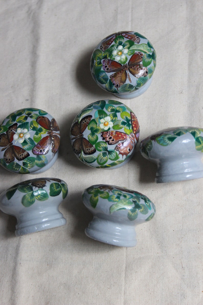 K&ouml;nnte beinhalten: Sechs Keramik-Schubladengriffe mit einer hellgrauen Basis und bemalten Designs von gr&uuml;nen Kleebl&auml;ttern, Schmetterlingen und wei&szlig;en Blumen.