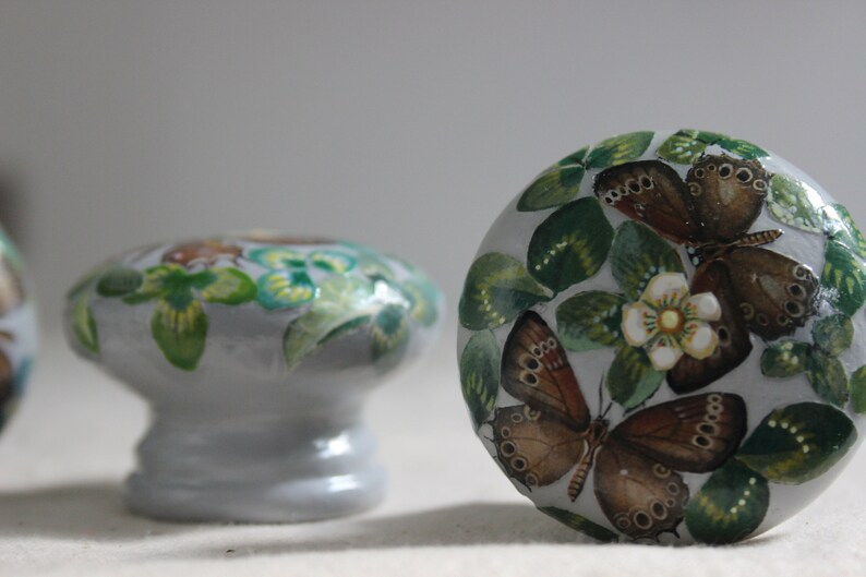 K&ouml;nnte beinhalten: Ein Satz von drei Keramik-Schubladengriffen mit einer hellgrauen Basis und handbemalten Schmetterlings- und Blumenmotiven. Die Schmetterlinge sind braun mit schwarzen Markierungen und die Blumen sind wei&szlig; mit gelben Zentren.