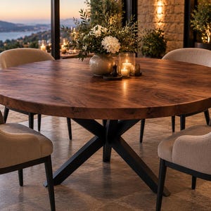 Puede incluir: Una mesa de comedor redonda grande con una superficie de madera de color marrón oscuro con vetas y una base de metal negro en forma de X. La mesa está rodeada por cuatro sillas tapizadas de tela beige claro con patas de madera oscura.