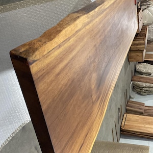 Puede incluir: Una tabla de madera larga y rectangular que presenta un borde natural en uno de sus lados. La madera muestra un tono marr&oacute;n medio c&aacute;lido con patrones de veta lineales visibles y un acabado de superficie liso y pulido.