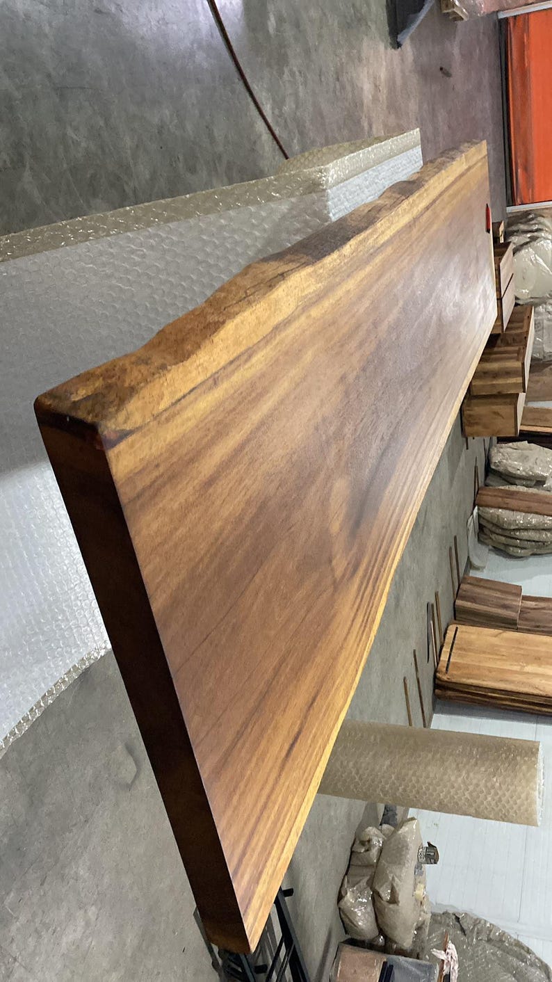Puede incluir: Una tabla de madera larga y rectangular que presenta un borde natural en uno de sus lados. La madera muestra un tono marr&oacute;n medio c&aacute;lido con patrones de veta lineales visibles y un acabado de superficie liso y pulido.