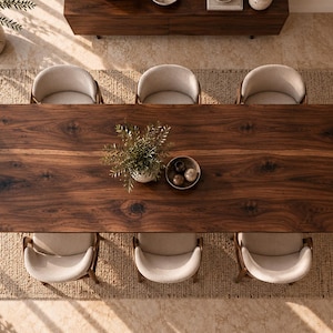 Puede incluir: Una mesa de comedor rectangular de madera marr&oacute;n oscuro con veta visible, rodeada de seis sillas tapizadas en color beige claro con respaldos curvos. Una peque&ntilde;a planta en maceta y un cuenco con tres esferas peque&ntilde;as descansan sobre la mesa.