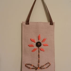 May include: A brown and white gingham fabric wall hanging with a brown ribbon loop for hanging. The fabric has an embroidered flower design with orange petals and a brown button center.