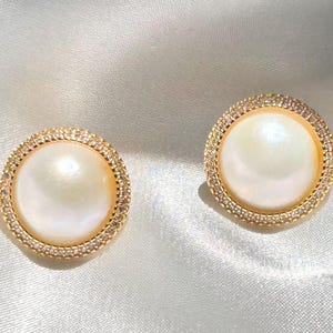 May include: A pair of round stud earrings featuring a central white iridescent pearl-like cabochon set in a gold-toned metal frame. The outer edge of each earring is lined with a circle of small, clear, sparkling stones.