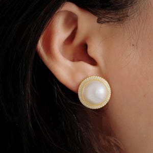 May include: A round stud earring featuring a large, smooth white pearl at the center, surrounded by a thin gold-toned border encrusted with small, clear sparkling stones. The circular design sits flush against the earlobe.