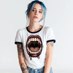 Puede incluir: Una camiseta blanca tipo ringer con ribetes negros en el cuello y las mangas, que presenta un estampado gráfico de una boca abierta con colmillos afilados y líquido rojo oscuro goteando de los labios, en un estilo ilustrativo detallado.