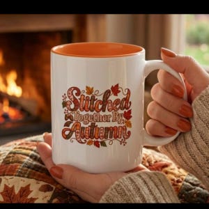May include: A white ceramic mug with an orange interior and handle, featuring the text Stitched Together By Autumn in brown and orange lettering, surrounded by colorful autumn leaf and acorn illustrations on the front surface.