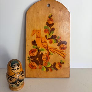 May include: A wooden wall hanging featuring an orange bird and floral design. The bird is on a branch with orange, brown, and green flowers. A Russian nesting doll is next to the wall hanging.