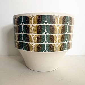 May include: A cream-colored ceramic planter with a flared base. The upper portion features a repeating pattern of green, brown, and gold geometric shapes. The planter is designed for indoor or outdoor use and adds a touch of vintage style.