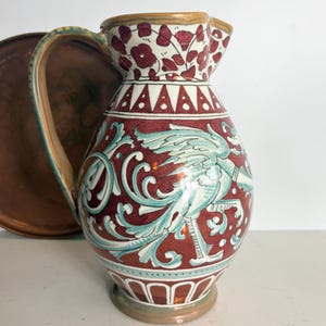May include: A decorative ceramic jug with a handle and spout. The jug features a maroon background with a detailed design of a bird and floral patterns in teal and white. The rim has a floral design.