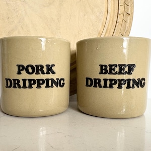 以下が含まれることがあります： 「PORK DRIPPING」と「BEEF DRIPPING」と黒い文字が書かれた2つのベージュ色の陶器の瓶。瓶は円筒形で、白い表面に置かれています。背景には木製のまな板があります。